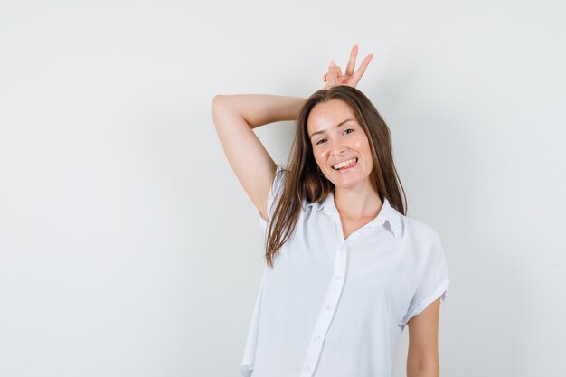 Young lady in white blouse making bunny gesture on her head and looking funny