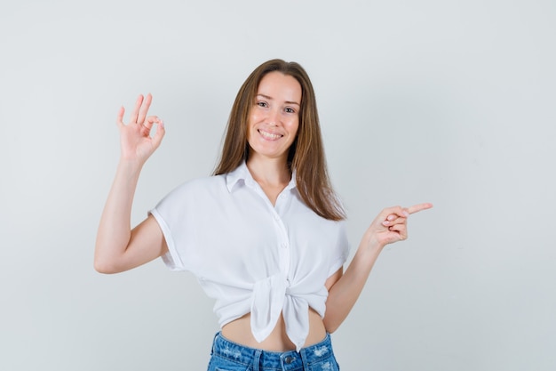 Young lady in white blouse showing ok gesture while pointing aside and looking positive , front view.