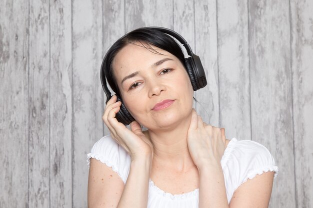 young lady in white shirt listening to music in black earphones on grey wall