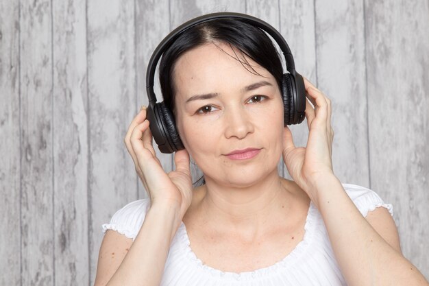 young lady in white shirt listening to music in black earphones on grey wall