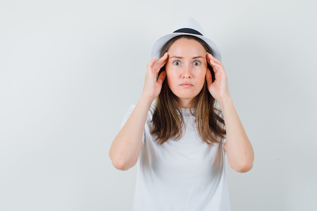 Free photo young lady in white t-shirt hat holding hands on temples and looking puzzled