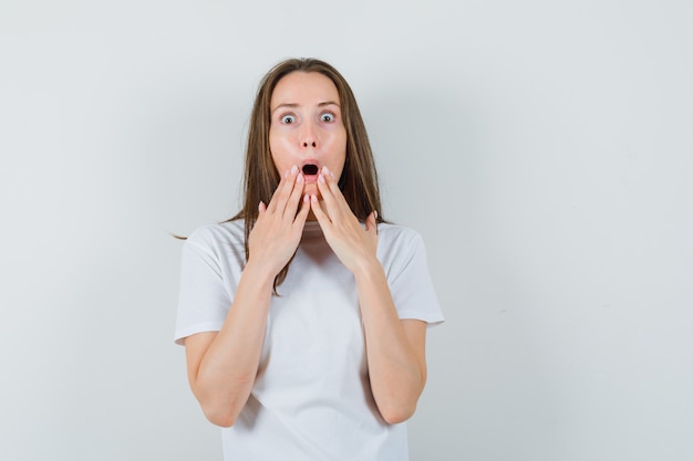 Young lady in white t-shirt holding hands near open mouth and looking surprised  