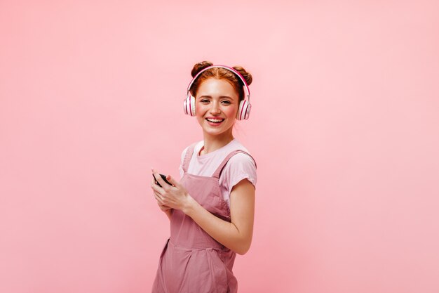 Young lady with buns looks at phone screen in surprise. Woman in dress and white T-shirt is listening to music with headphones on pink background.