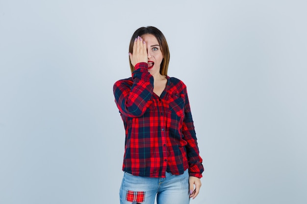 Young lady with hand on eye, opening mouth in checked shirt, jeans and looking astonished. front view.