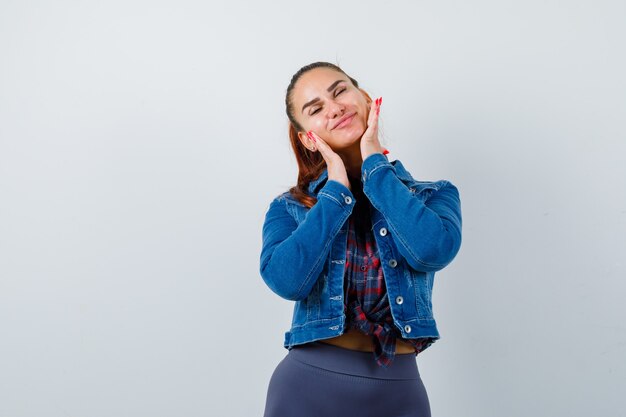 Young lady with hands on cheeks in checkered shirt, denim jacket and looking peaceful , front view.