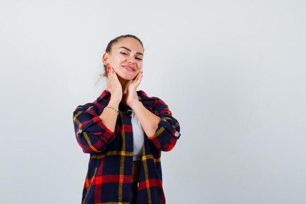 Young lady with hands on chin in top, plaid shirt and looking joyful , front view.