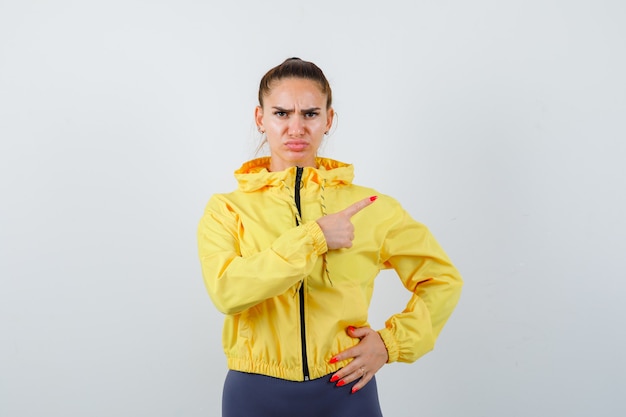 Free photo young lady in yellow jacket pointing at upper right corner and looking serious , front view.