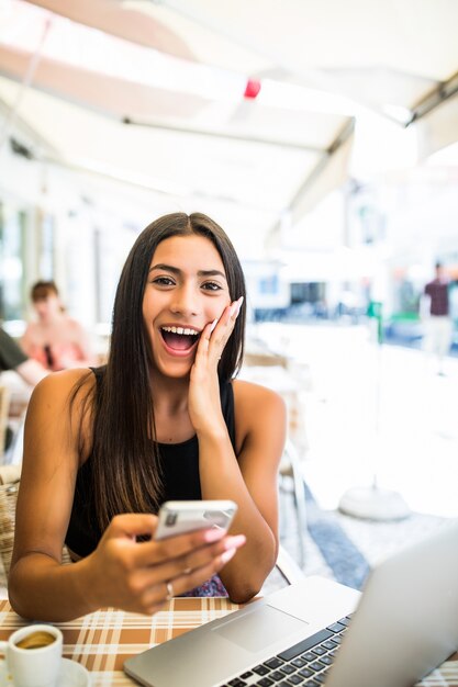 Young latin woman getting shocked by news in her phone outdoors in cafe. Shocked face of a girl sitting at a terrace with her phone in hands.