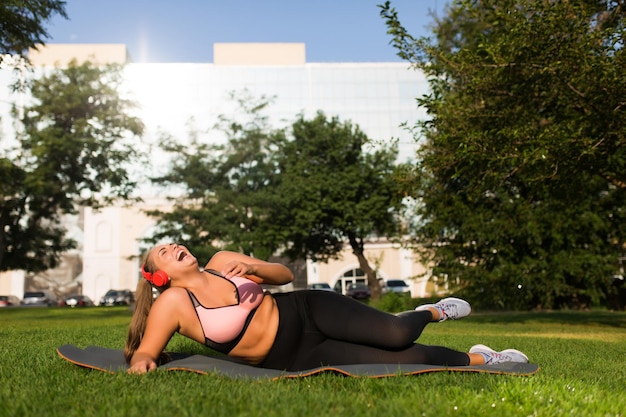 Free photo young laughing plus size woman in sporty top and leggings with red headphones lying on yoga mat while happily spending time in city park