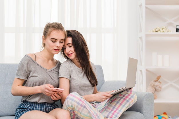 Young lesbian holding laptop on her lap looking in girlfriend's mobile