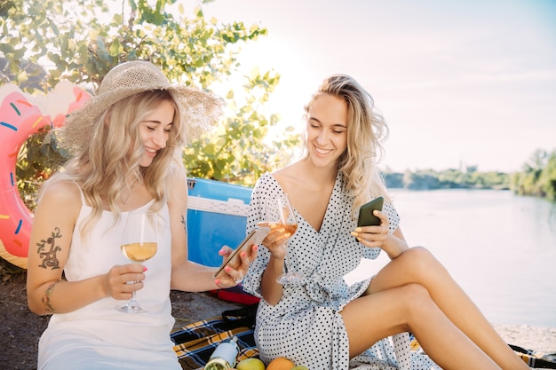 Free Photo young lesbian's couple having fun at riverside in sunny day. women spending time on the nature together. drinking wine, making selfie. concept of relationship, love, summer, weekend, honeymoon.