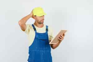 Free photo young loader holding board with hand on head in uniform and looking pensive , front view.