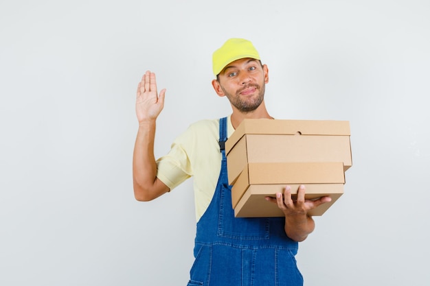 Young loader in uniform holding cardboard boxes and saying hello, front view.