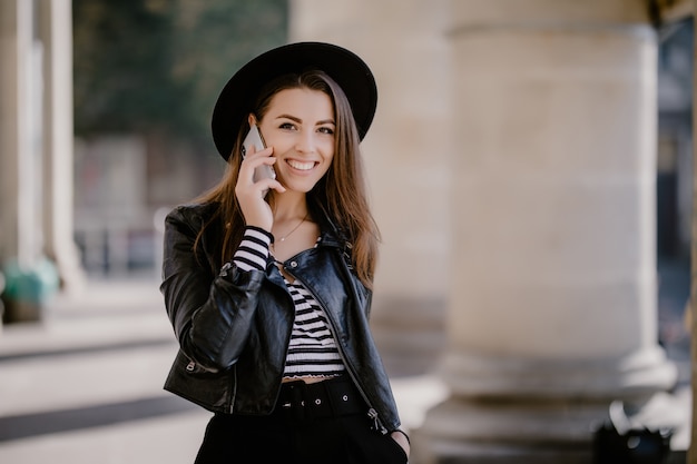 Young lovely brown-haired girl in a leather jacket, black hat on the city promenade have a phone conversation