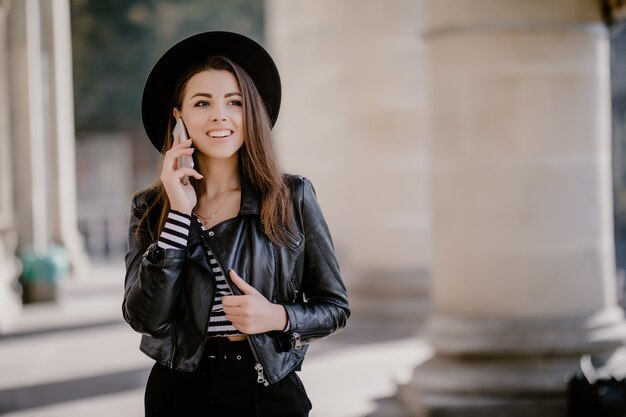 Young lovely brown-haired girl in a leather jacket, black hat on the city promenade have a phone conversation