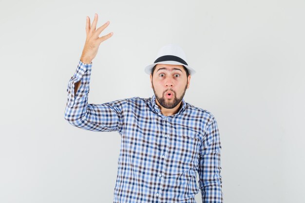 Young male in checked shirt, hat raising arm in surprised gesture , front view.