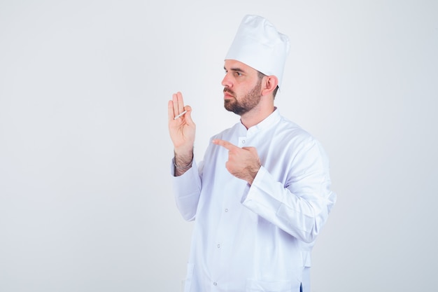 Young male chef pointing at cigarette in white uniform and looking thoughtful. front view.