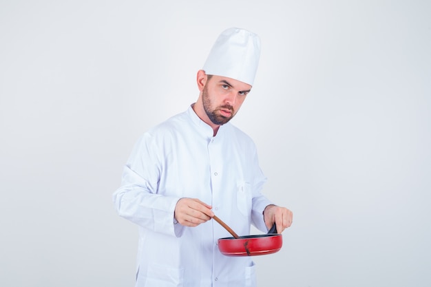 Free photo young male chef in white uniform mixing meal with wooden spoon and looking pensive , front view.