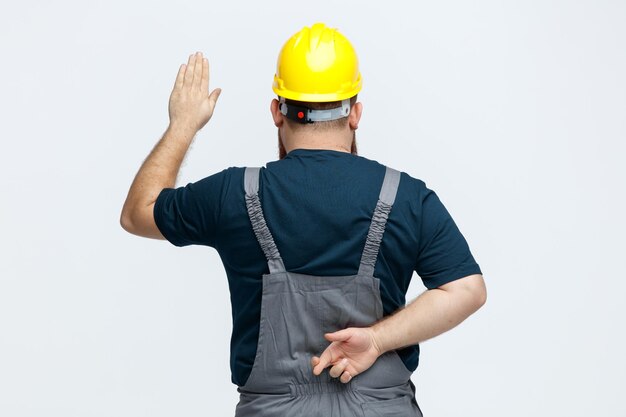 Young male construction worker wearing safety helmet and uniform standing in behind view showing stop gesture while crossing fingers behind his back isolated on white background