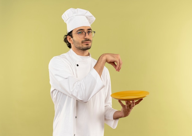 young male cook wearing chef uniform and glasses holding plate and pretending spilling salt 