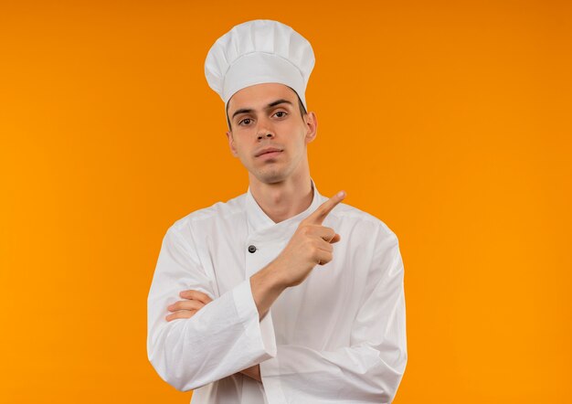  young male cool wearing chef uniform points finger to side on isolated yellow wall with copy space