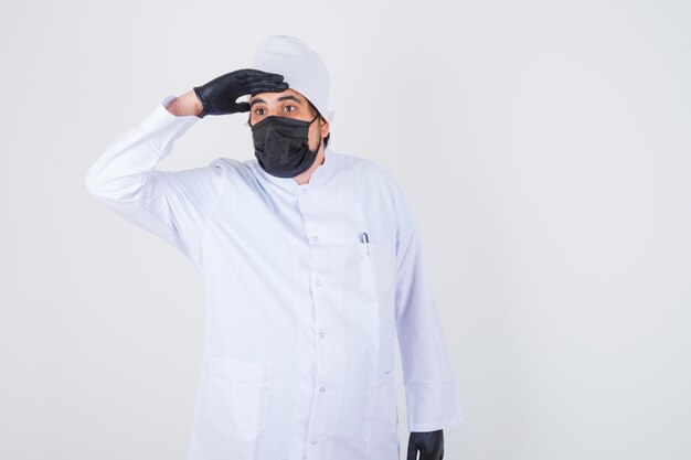 Young male doctor holding hand on forehead while looking away in white uniform and looking focused. front view.