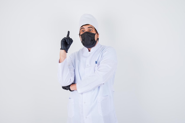 Young male doctor pointing up in white uniform and looking confident , front view.