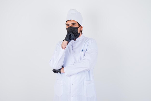 Young male doctor propping chin on fist in white uniform and looking thoughtful. front view.