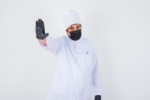 Young male doctor showing stop gesture in white uniform and looking confident. front view.