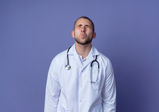 Young male doctor wearing medical robe and stethoscope around his neck doing kiss gesture with closed eyes isolated on purple background with copy space