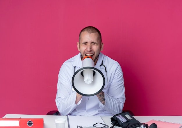 Young male doctor wearing medical robe and stethoscope sitting at desk with work tools shouting in loud speaker with closed eyes isolated on pink background