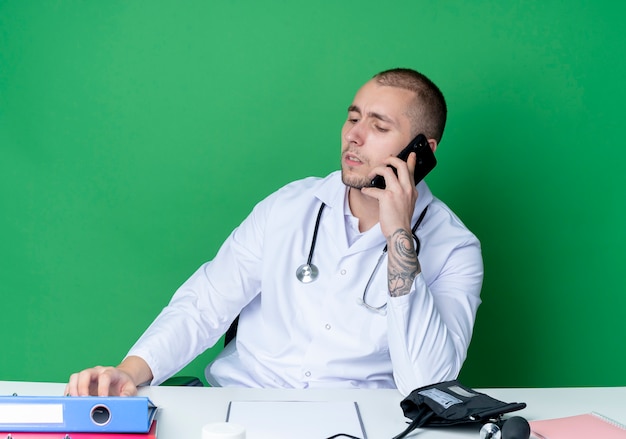 Free Photo young male doctor wearing medical robe and stethoscope sitting at desk with work tools talking on phone and looking down with hand on desk isolated on green background