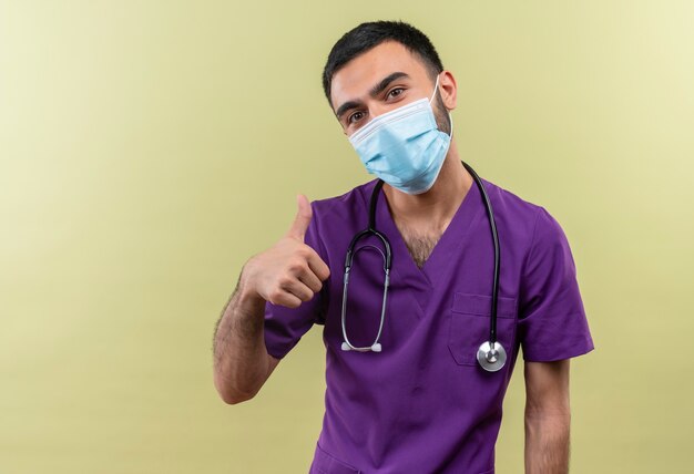 young male doctor wearing purple surgeon clothing and stethoscope medical mask his thumb up on isolated green wall