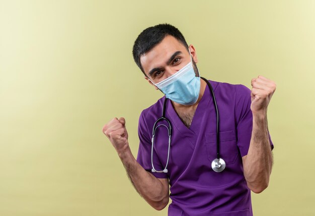 young male doctor wearing purple surgeon clothing and stethoscope medical mask showing yes gesture on isolated green wall