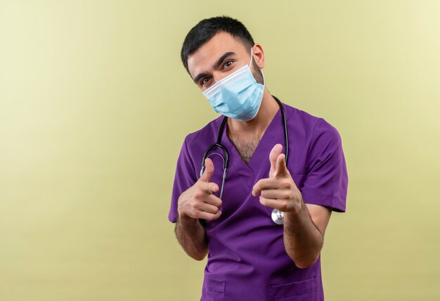 young male doctor wearing purple surgeon clothing and stethoscope medical mask showing you gesture on isolated green wall