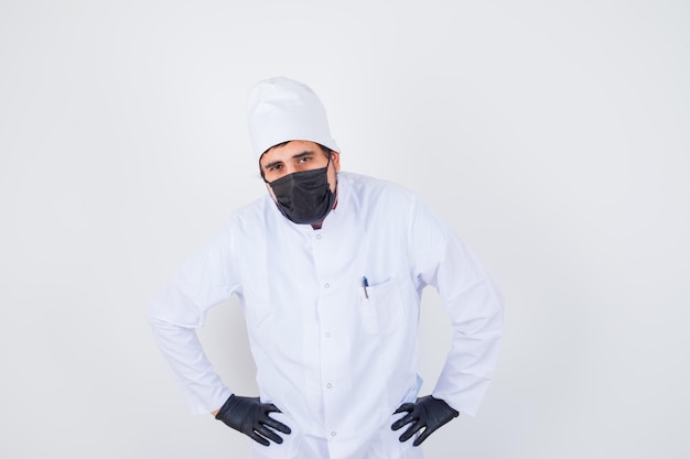 Young male doctor in white uniform holding hands on waist and looking fatigued , front view.