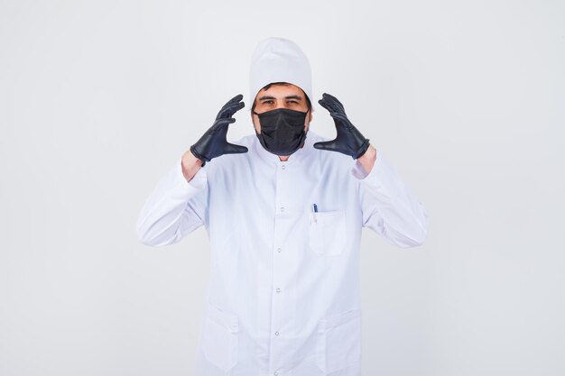 Young male doctor in white uniform raising hands in aggressive manner and looking annoyed , front view.