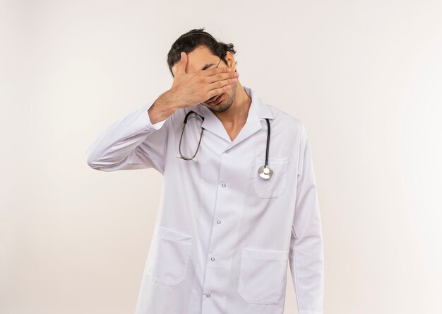 Young male doctor with optical glasses wearing white robe with stethoscope covered eyes with hand on isolated white wall with copy space