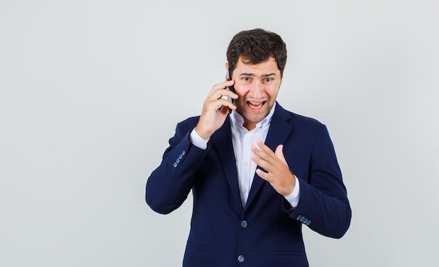 Young male getting angry while talking on smartphone in suit , front view.