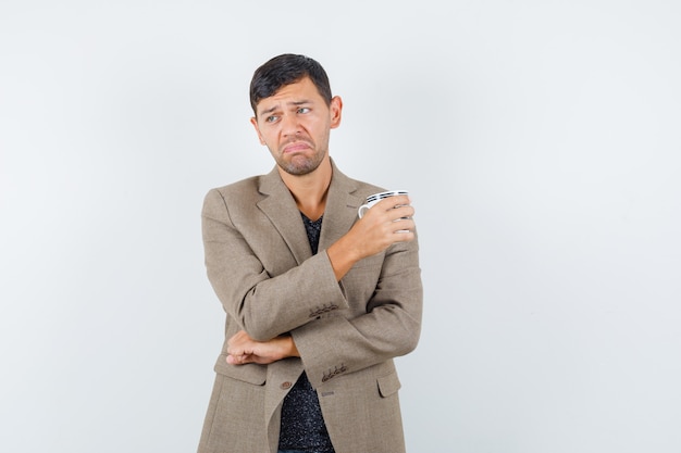 Young male in grayish brown jacket holding cup and looking displeased , front view.