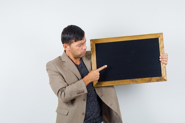 Young male in grayish brown jacket pointing to cardboard and looking thoughtful , front view.