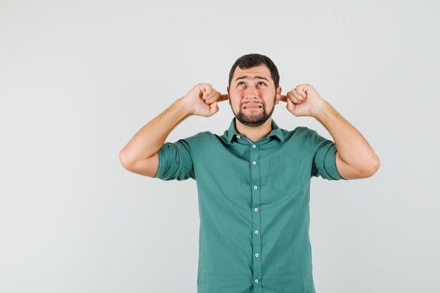 Young male in green shirt plugging ears with fingers and looking scared , front view.