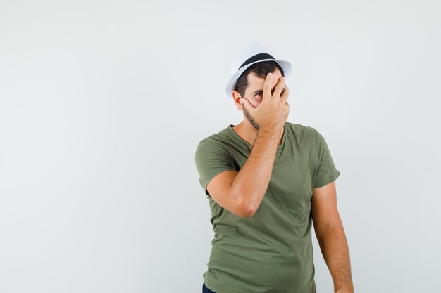 Young male in green t-shirt and hat holding hand on face and looking scared, front view.