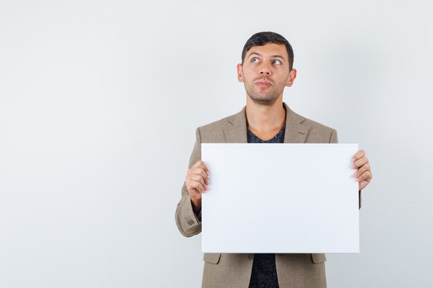 Young male holding blank paper in grayish brown jacket and looking thoughtful. front view.