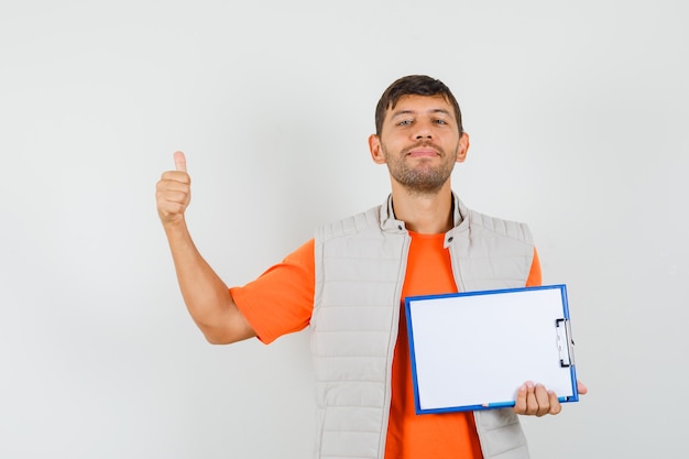 Free Photo young male holding clipboard, pencil, showing thumb up in t-shirt, jacket and looking jolly , front view.