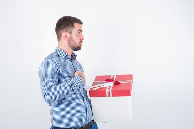 Young male holding gift box while keeping hand on chest in shirt and looking perplexed , front view.