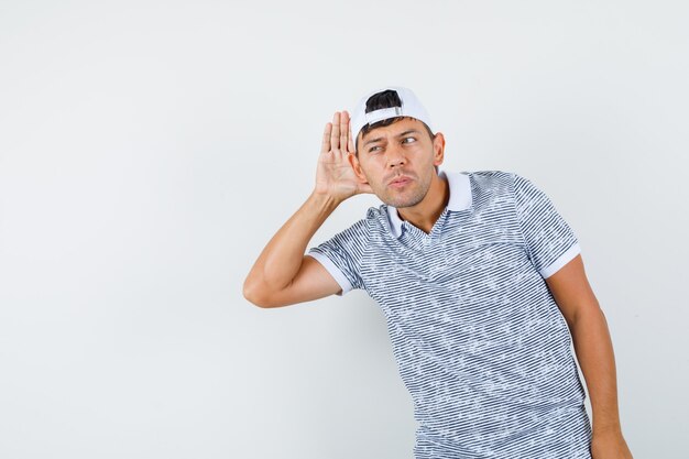 Young male holding hand behind ear to listen in t-shirt and cap and looking curious 