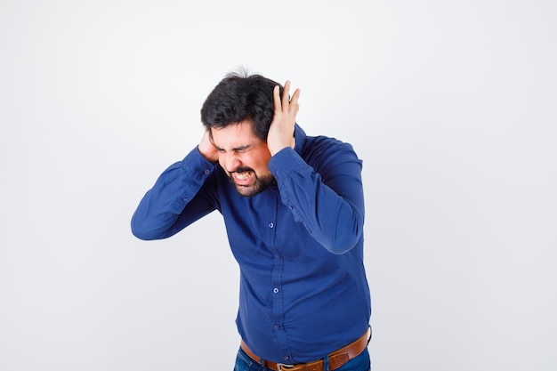 Young male holding hand on ears while shouting in royal blue shirt and looking angry. front view.