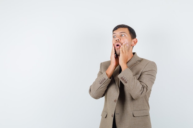 Young male holding hands on his face in grayish brown jacket and looking confused. front view.