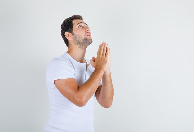 Young male holding hands in praying gesture in white t-shirt and looking hopeful